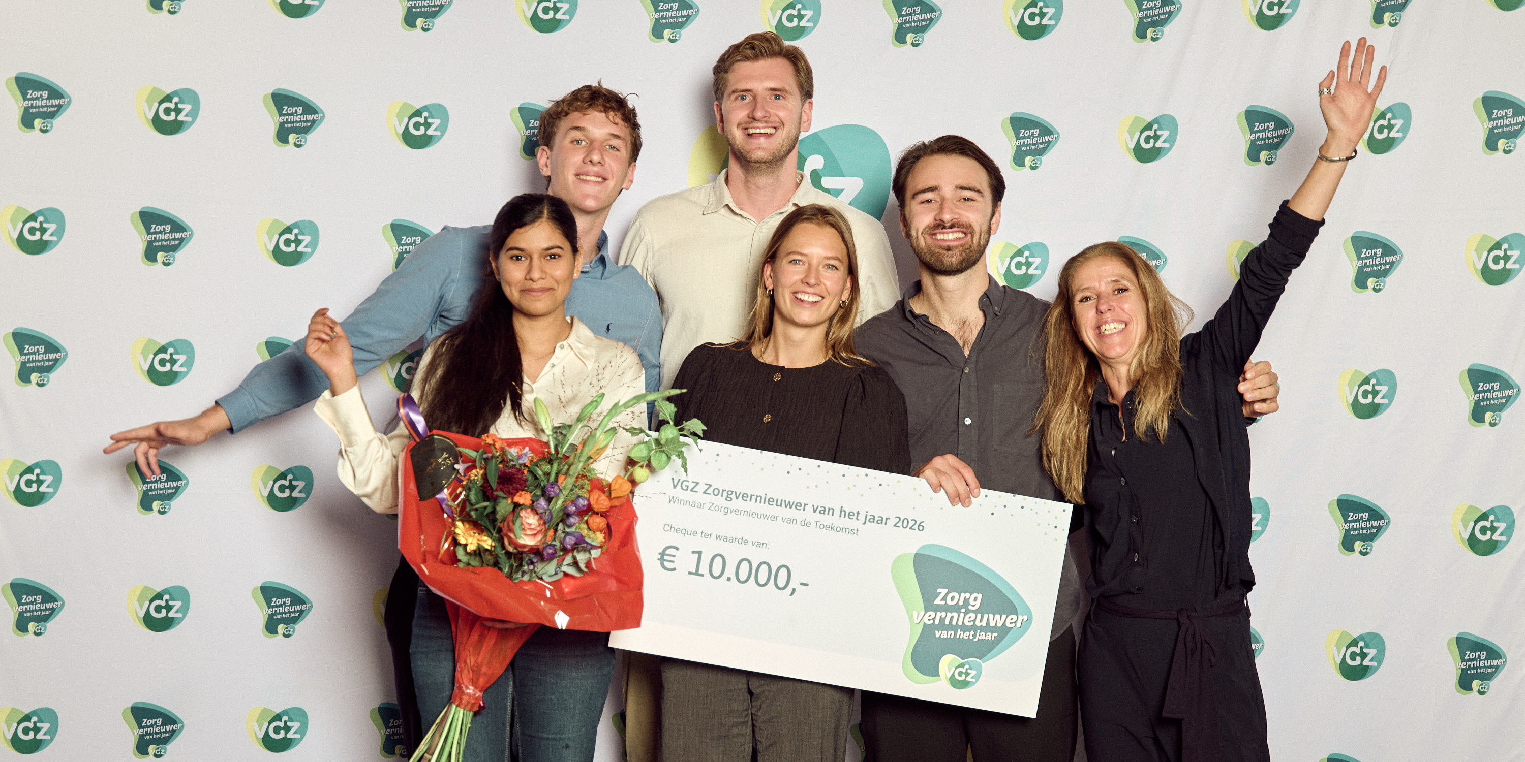 Het PAVA-team voor de fotowand van VGZ met de cheque van 10.000 euro voor Winnaar Zorgvernieuwer van de Toekomst, met bloemen en een arm in de lucht